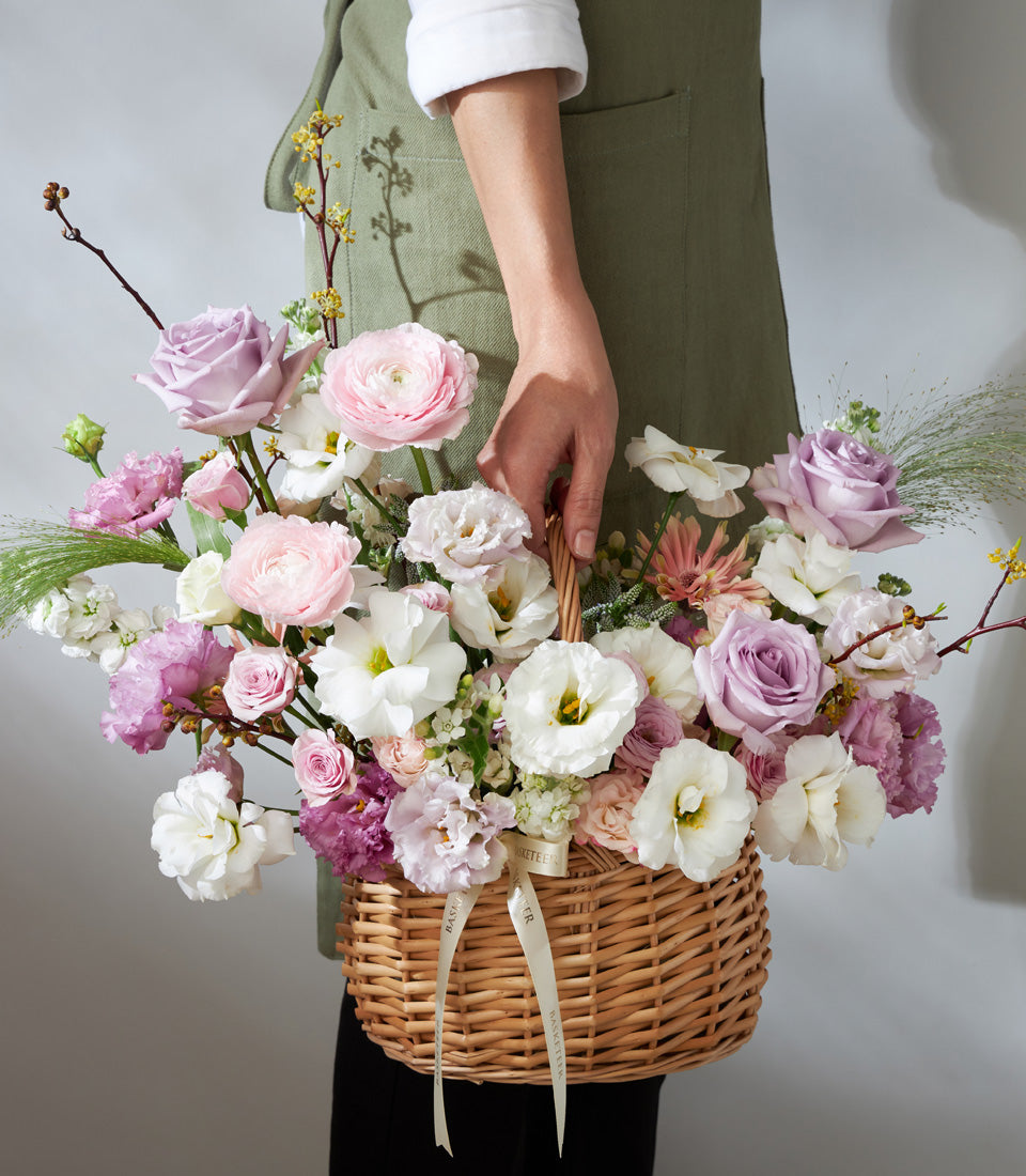 Woven wicker basket of lavender and pink roses, ranunculus, and lisianthus with cream ribbons