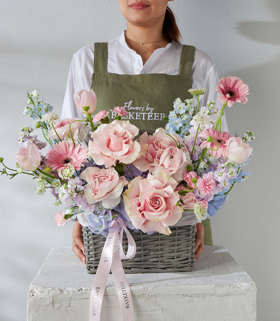 Pastel pink roses and gerbera daisies in grey wicker basket with light blue delphiniums and ribbon