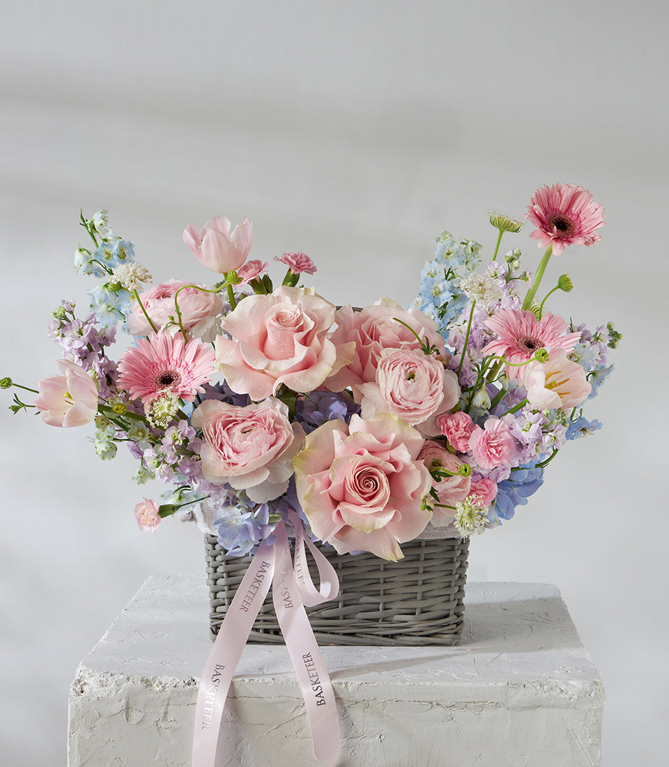 Pastel roses and pink gerbera daisies in grey woven basket with blue delphiniums