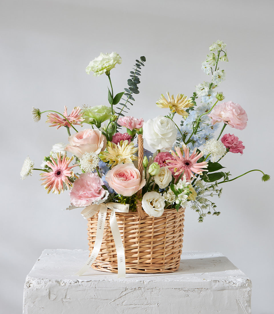 Pastel Bloom flower basket with pink roses, white gerberas, and blue delphiniums in wicker for birthday gifts