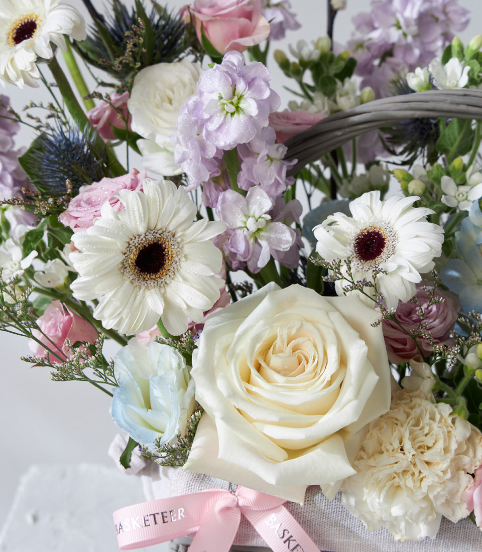 Pastel Serenity flower basket with white gerbera daisies, pink roses, and purple stock in grey wicker