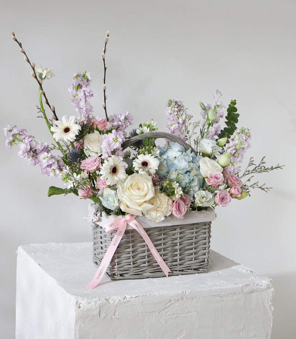 Pastel flower basket with white roses, blue hydrangeas, pink spray roses, and purple stock arranged in grey wicker