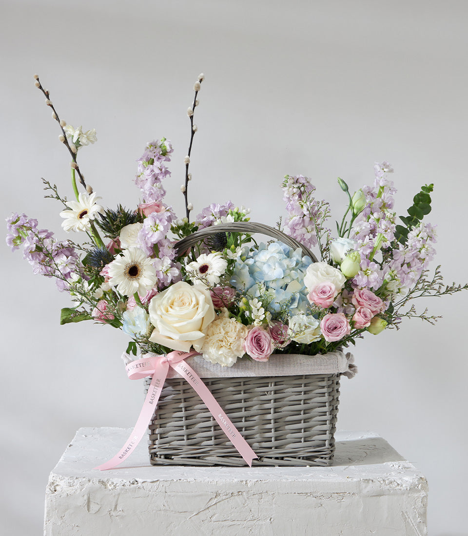 Pastel pink and white roses with hydrangeas in grey wicker basket with satin ribbon