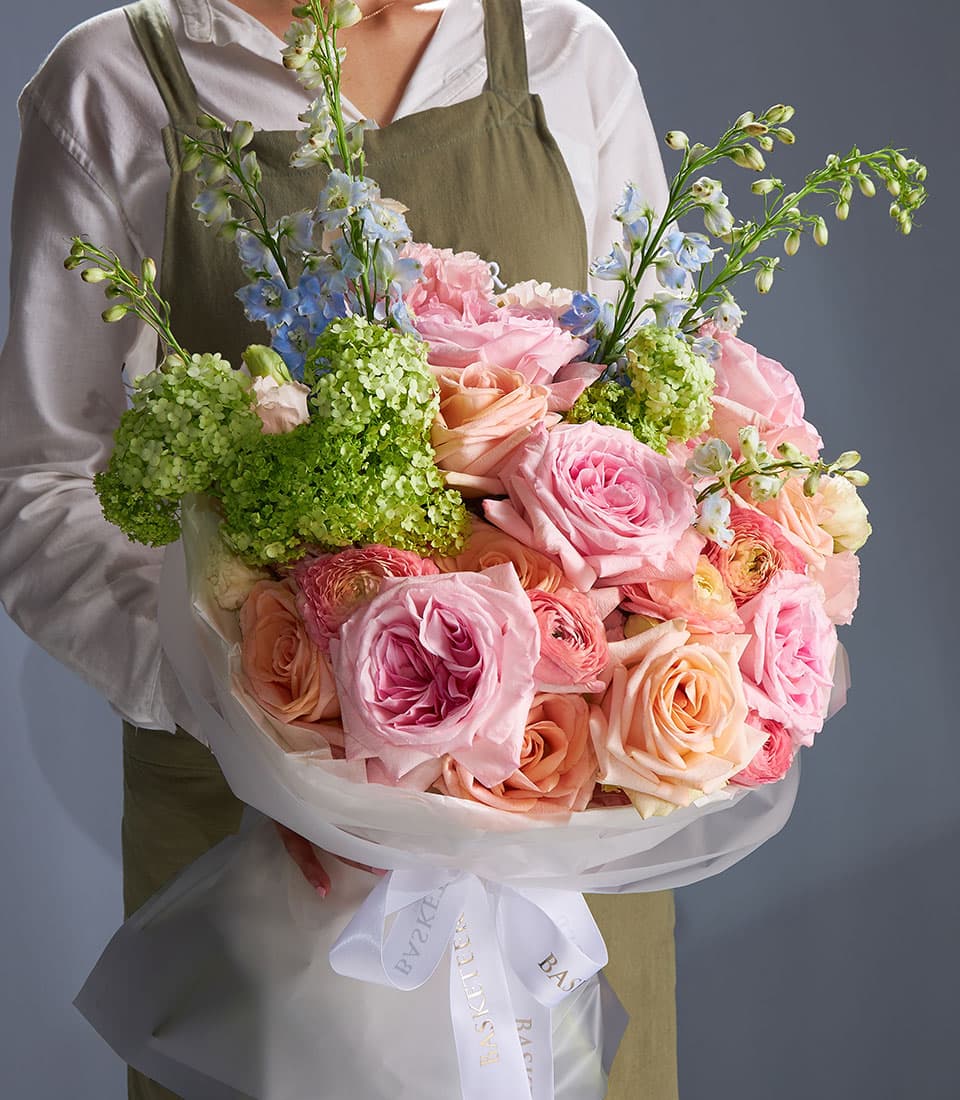 Pastel rose and ranunculus bouquet in pink, peach, and cream with hydrangea and delphinium, wrapped in white cellophane