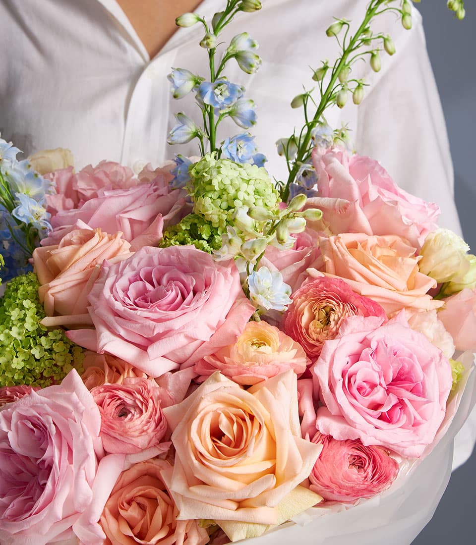 Pastel pink, peach and coral rose bouquet with blue delphiniums and green filler flowers, wrapped in clear cellophane