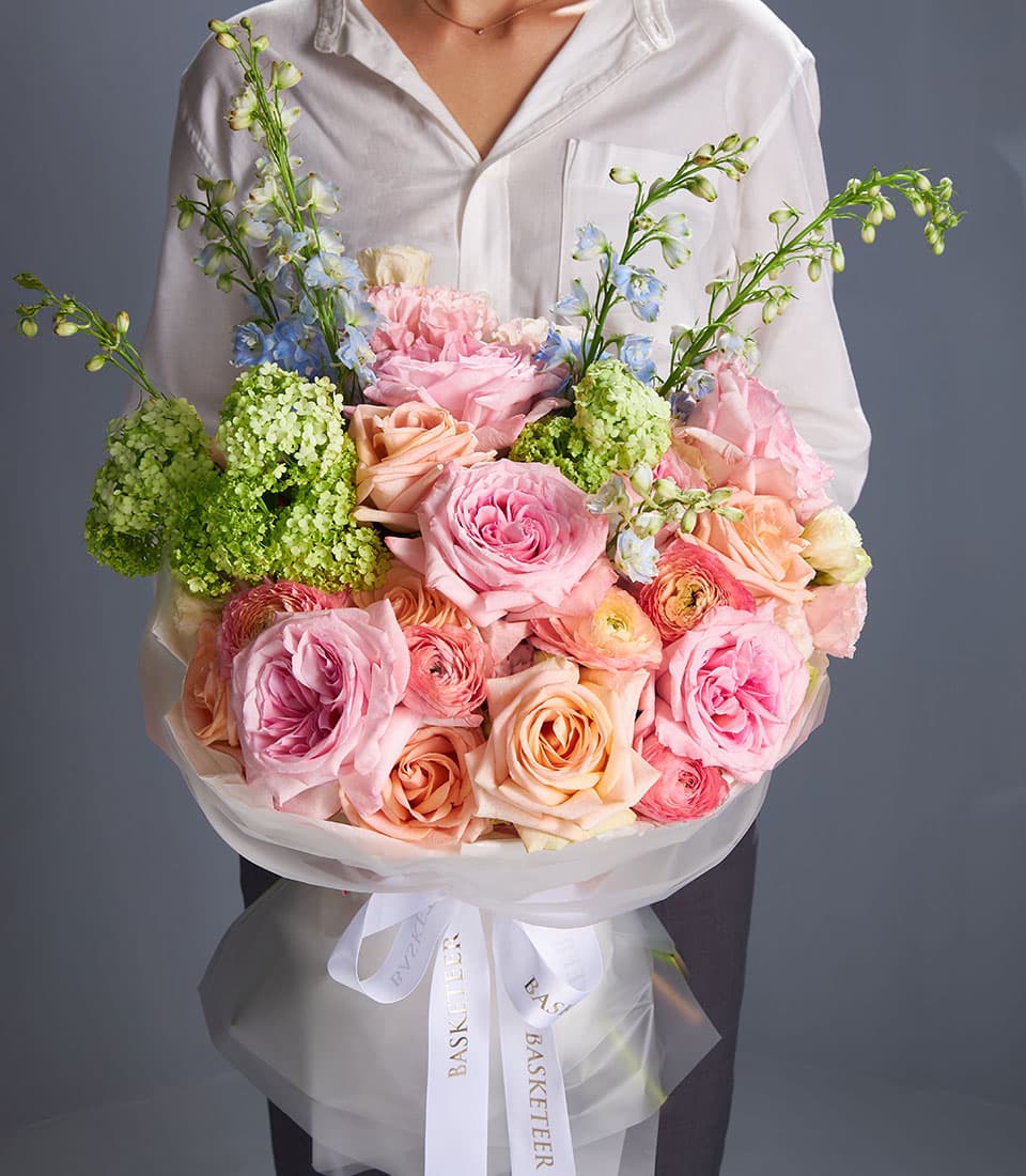 Pastel O'Hara and Tiffany Rose bouquet with pink roses, ranunculus, hydrangeas, and delphinium in white paper wrapping