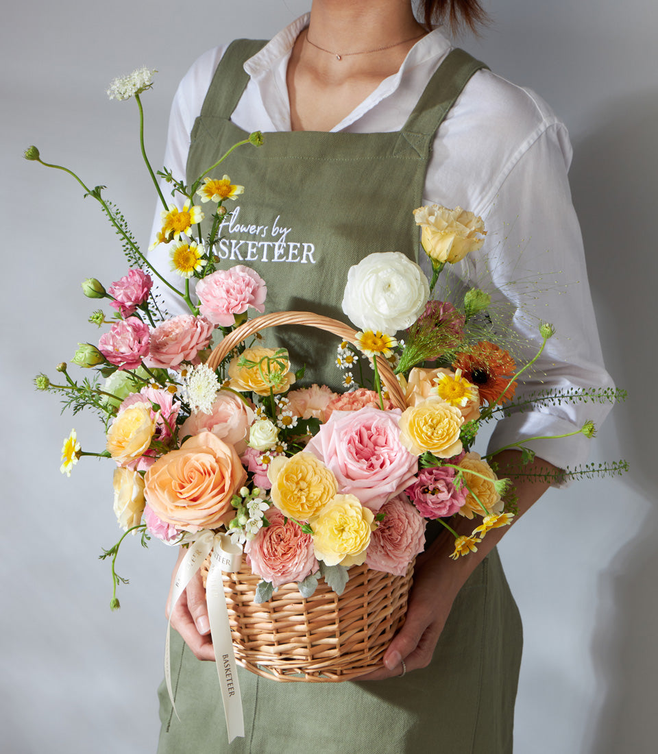 Wicker basket of fresh peach, pink, and yellow roses with ranunculus and daisies, birthday flower gift