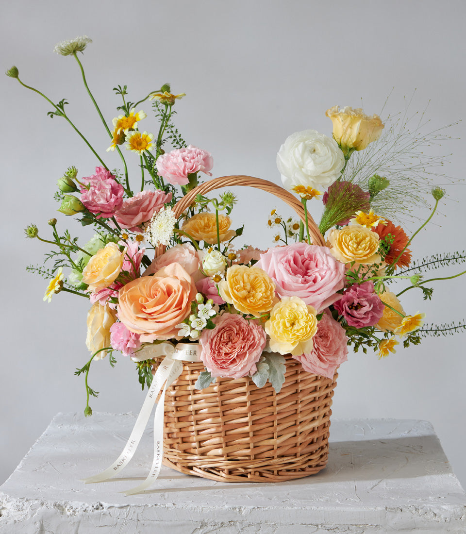 Radiant pink and peach roses with ranunculus in a woven wicker basket, tied with cream ribbon for birthday celebrations