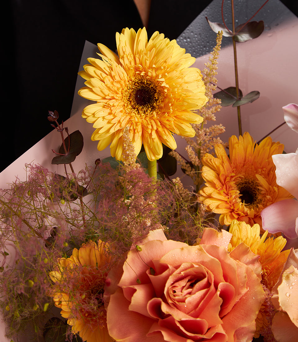 Vibrant fresh flower bouquet with yellow and orange gerberas, peach rose, and pink smoke bush in two-toned wrapping