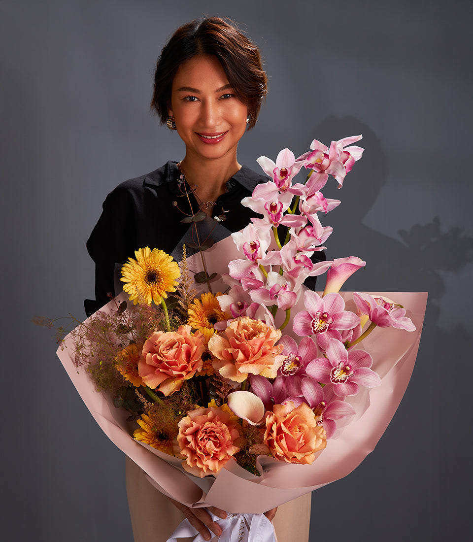Woman holding vibrant luxury bouquet with yellow gerberas, orange roses, and pink orchids wrapped in blush paper