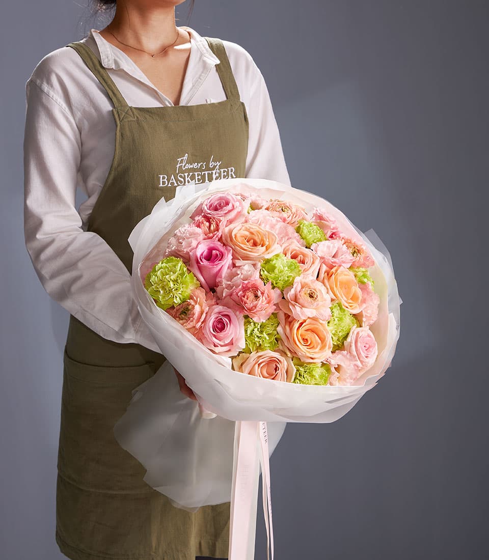Florist in khaki apron holding Blooming Delight mixed bouquet with pink roses, peach roses, and light green carnations in