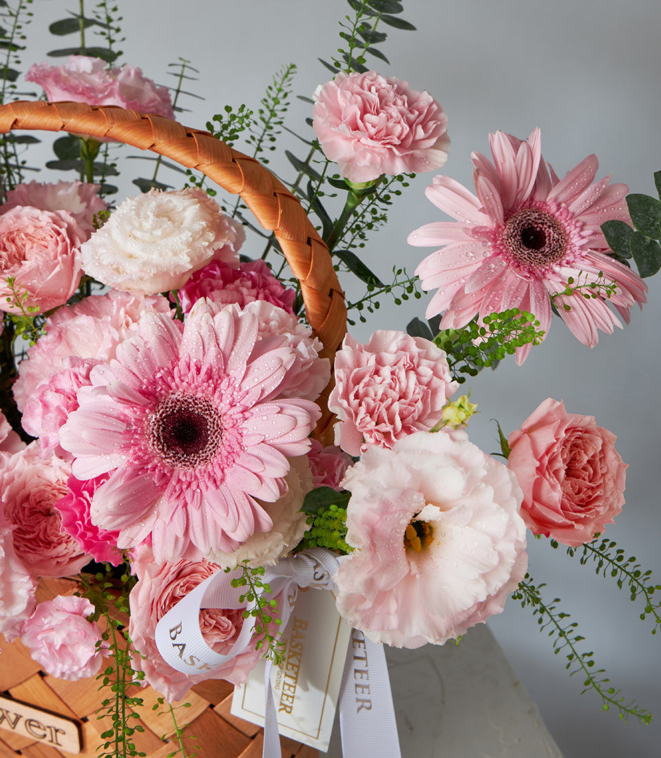 Vibrant pink gerbera daisies, carnations, and roses in a woven basket with white ribbon, perfect for birthday celebrations