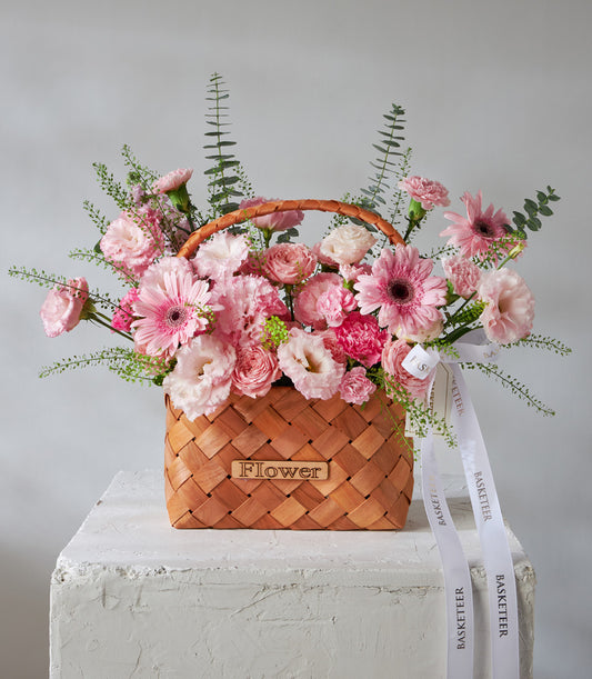 Pink gerbera daisies, roses, and lisianthus in woven basket with green foliage, birthday flower gift