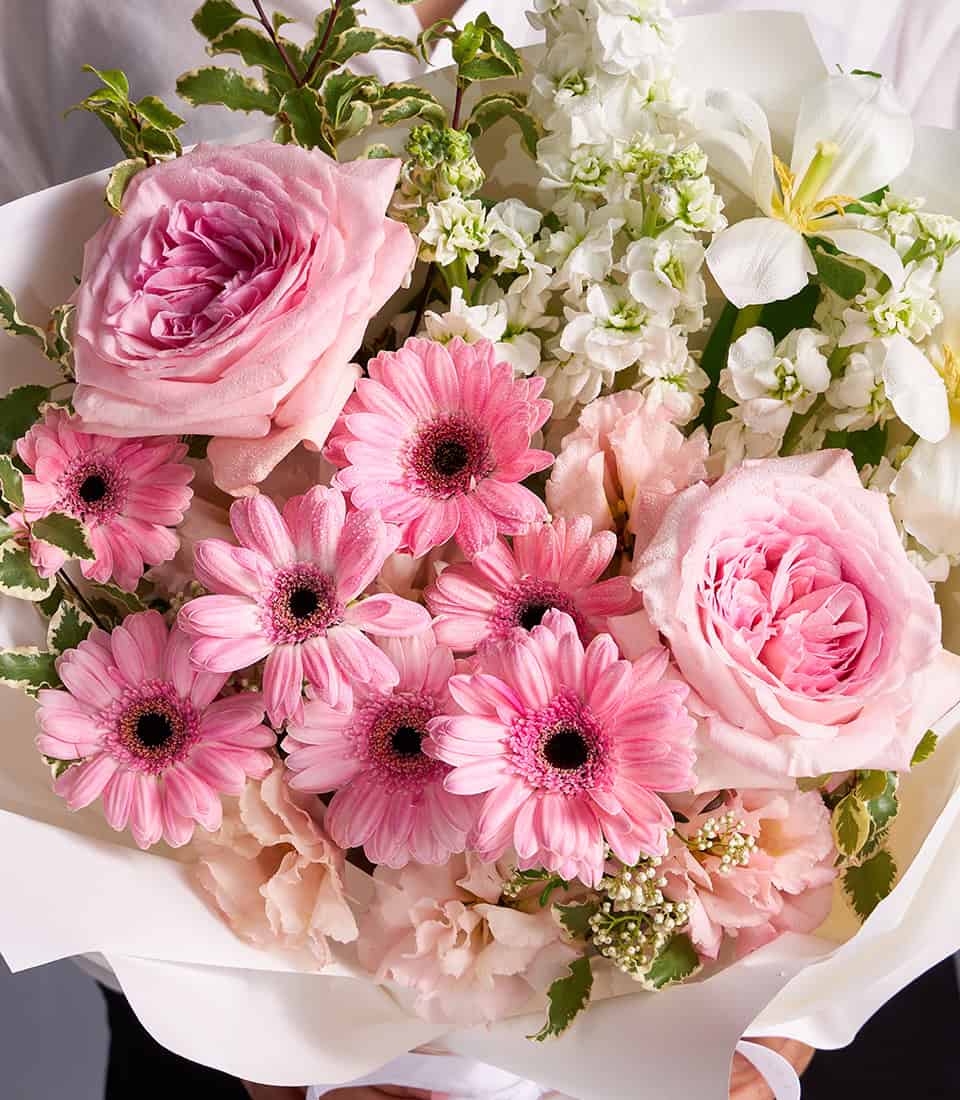 Pink garden roses, gerberas, and white stock bouquet with fresh green foliage, hand-tied in white paper