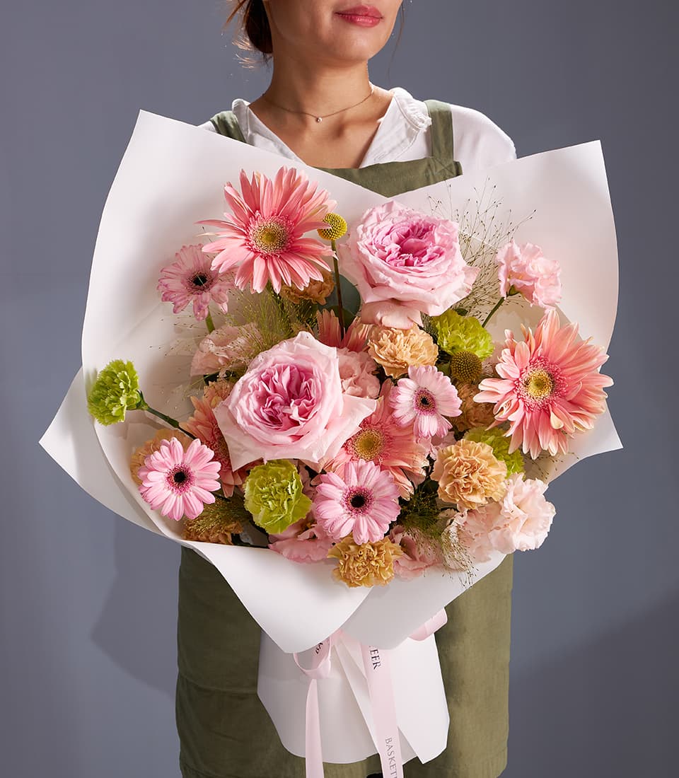 Pastel bouquet of pink garden roses, gerbera daisies, and peach carnations wrapped in white paper with pink ribbon