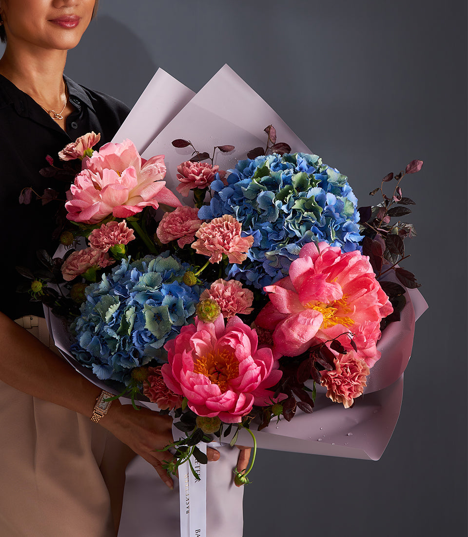 Luxury bouquet of pink peonies, blue hydrangeas, and carnations wrapped in light purple paper with white ribbon