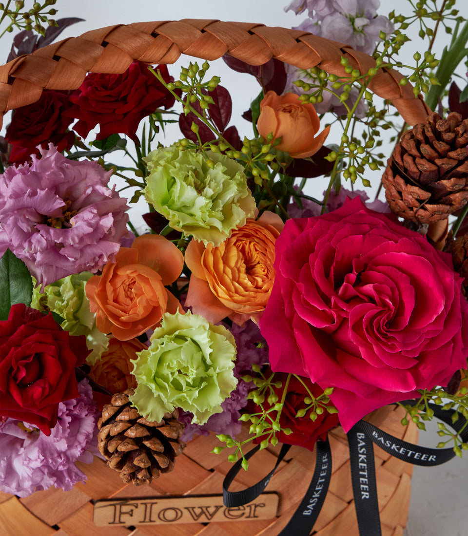 Crimson and fuchsia rose arrangement in woven basket with pinecones and greenery, artisan birthday flowers