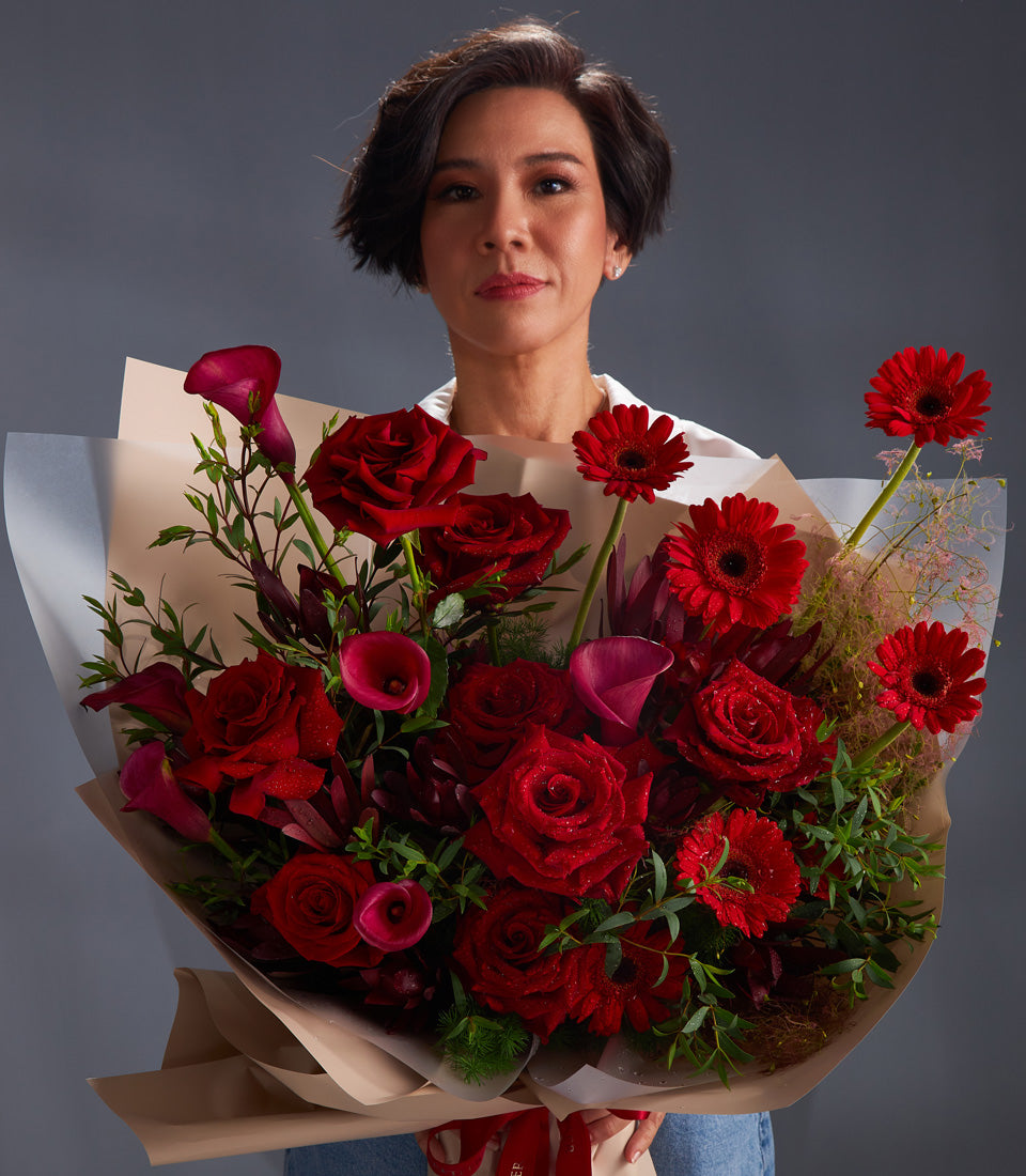 Woman holding lush red rose, gerbera daisy, and calla lily bouquet wrapped in cream paper with red ribbon