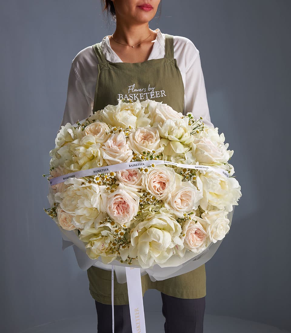 Florist holding ivory and blush rose bouquet with white filler flowers and green foliage, wrapped in sheer white with