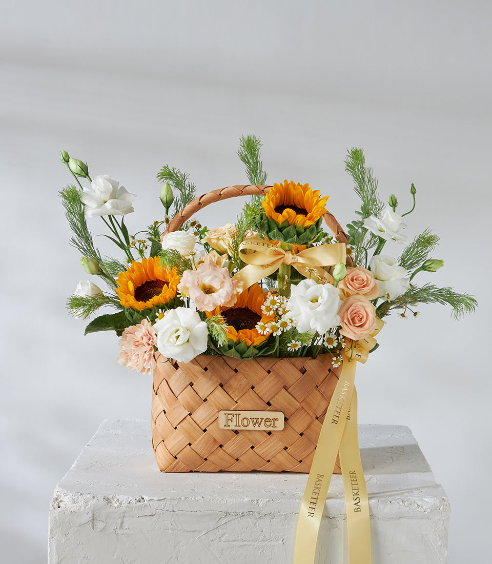 Woven wood basket filled with bright yellow sunflowers, white roses, and peach spray roses tied with golden ribbon