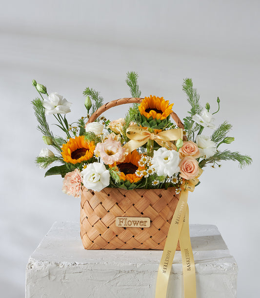 Woven wood basket filled with bright yellow sunflowers, white roses, and peach spray roses tied with golden ribbon