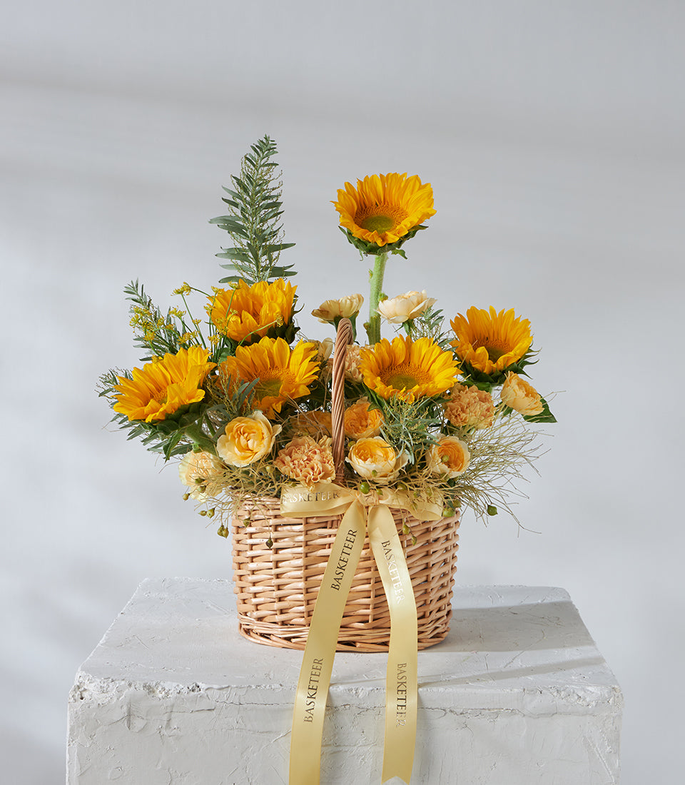 Vibrant sunflowers and yellow roses in woven wicker basket with green foliage and gold ribbon