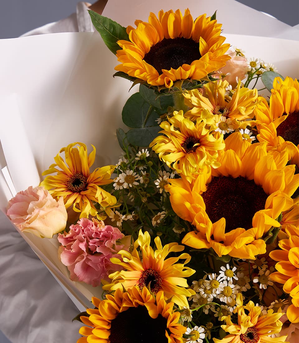 Vibrant sunflower and daisy garden bouquet with yellow blooms, white fillers, and soft pink accents in cream wrapping