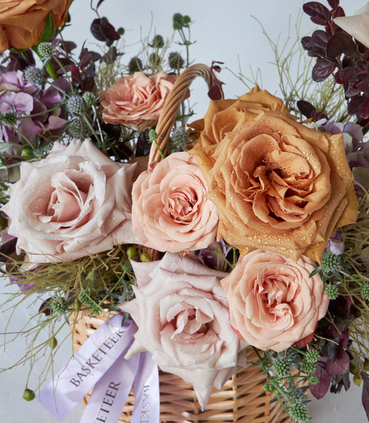 Woven wicker basket with peach, pink, and coral roses, purple hydrangea, and thistle greenery arrangement