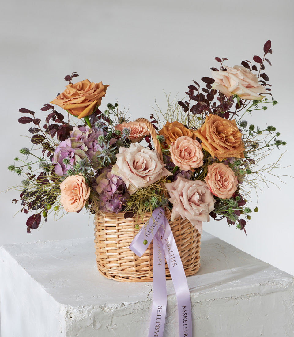 Peach and pink roses with purple hydrangeas in woven wicker basket, luxury gift arrangement