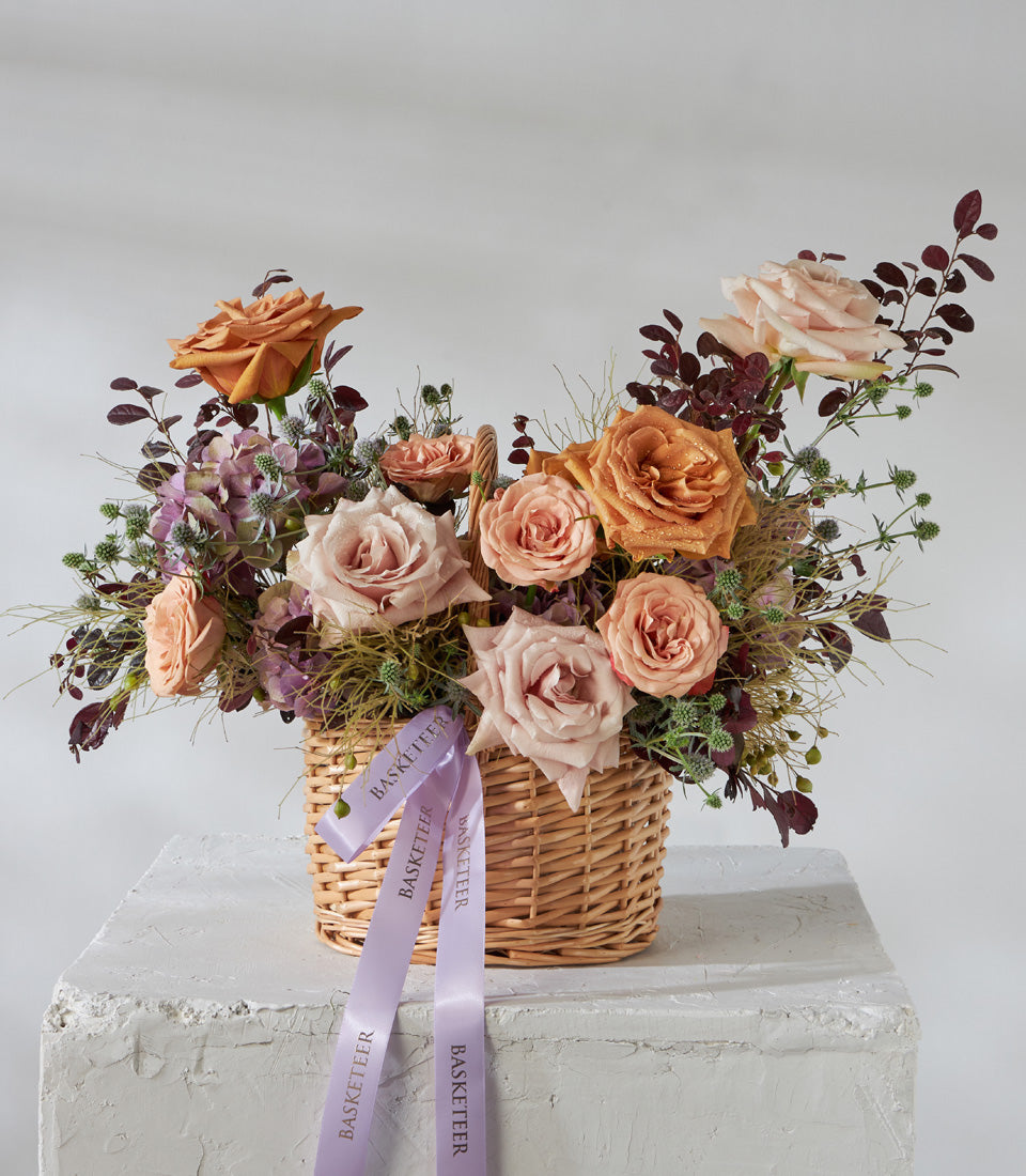 Wicker basket with peach, orange, and pink roses, purple hydrangeas, and green thistles tied with purple Basketeer ribbons