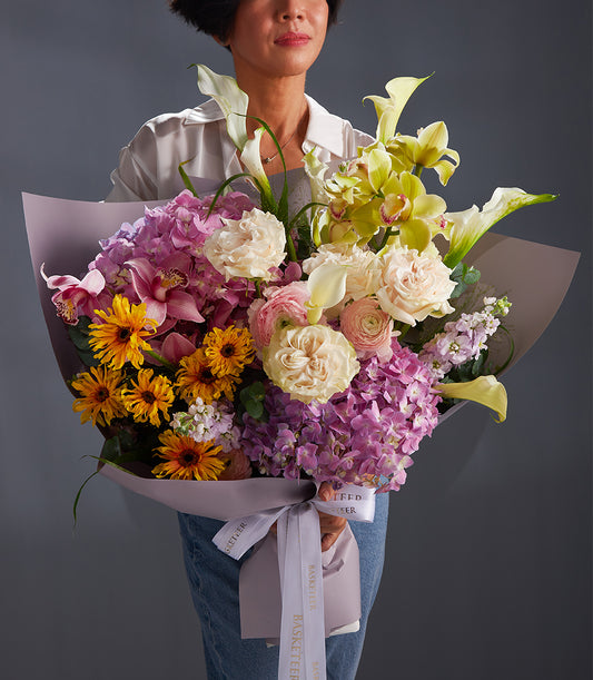 Vibrant mixed flower bouquet with pink hydrangeas, sunflowers, roses, orchids, and calla lilies in lavender paper