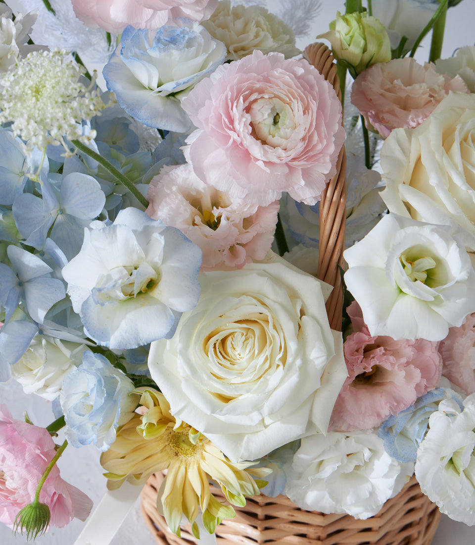 Pastel Radiance wicker flower basket with white roses, pink ranunculus, blue hydrangeas, and yellow gerberas for birthdays