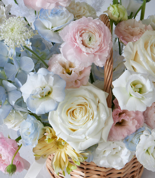Pastel Radiance wicker flower basket with white roses, pink ranunculus, blue hydrangeas, and yellow gerberas for birthdays