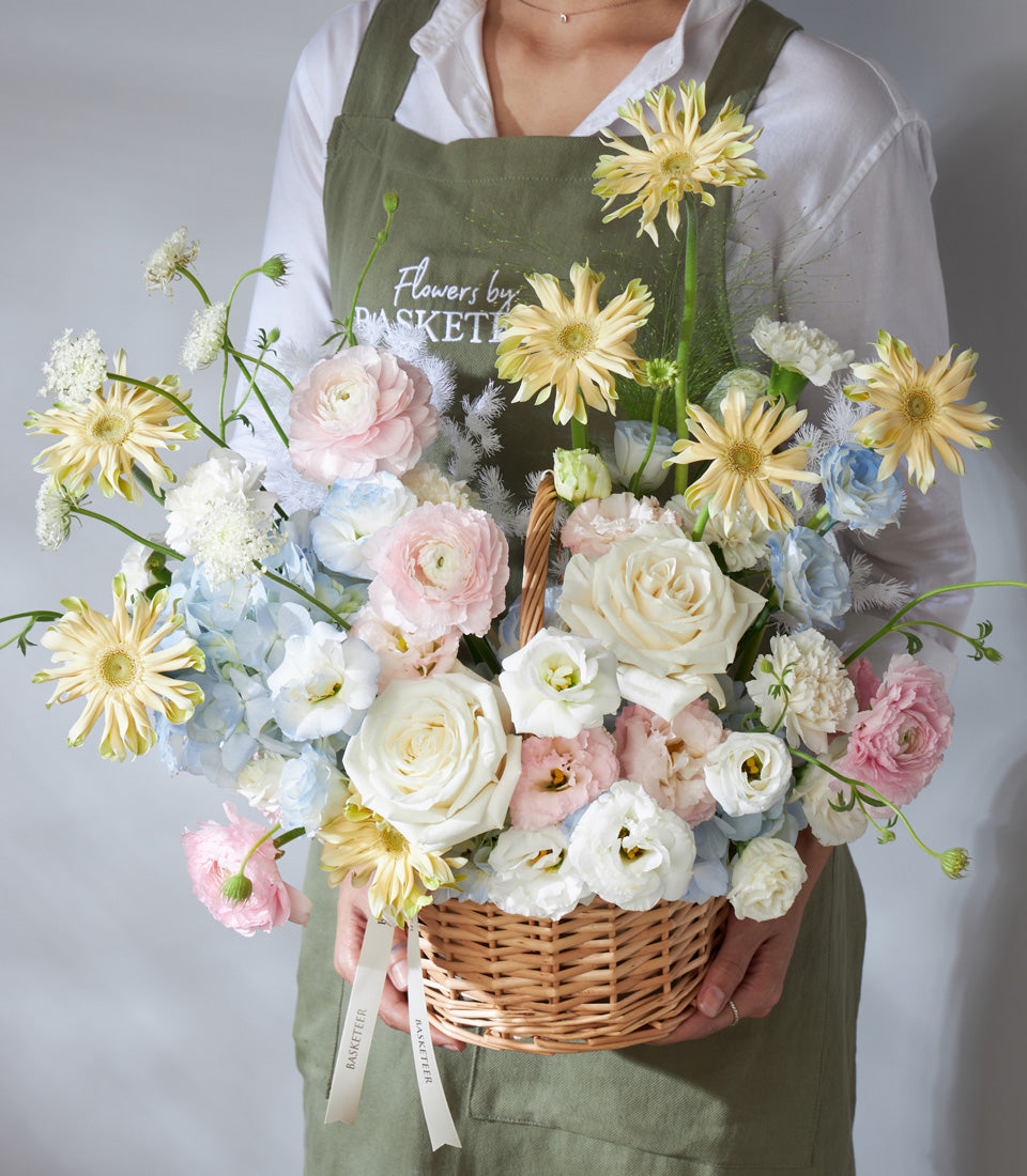 Pastel Radiance wicker flower basket with white, blush pink, light blue, and pale yellow ranunculus, roses, hydrangeas, and