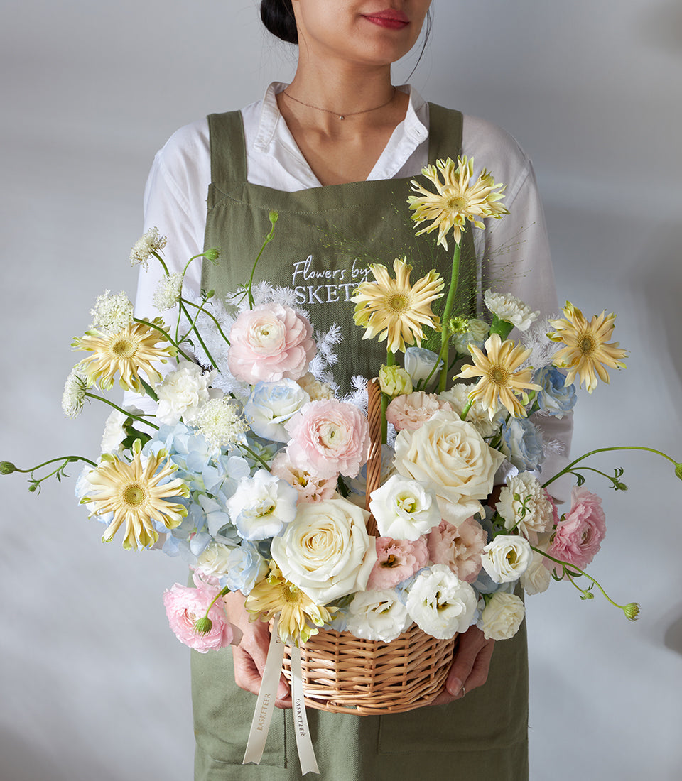 Florist holding large wicker basket of pastel pink ranunculus, white roses, blue hydrangeas, and yellow gerberas for