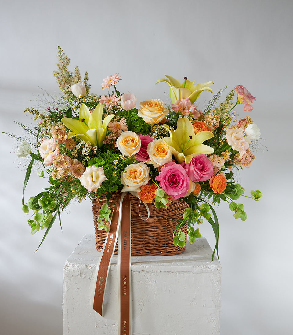 Pastel yellow lilies and pink roses in woven basket with white tulips and greenery