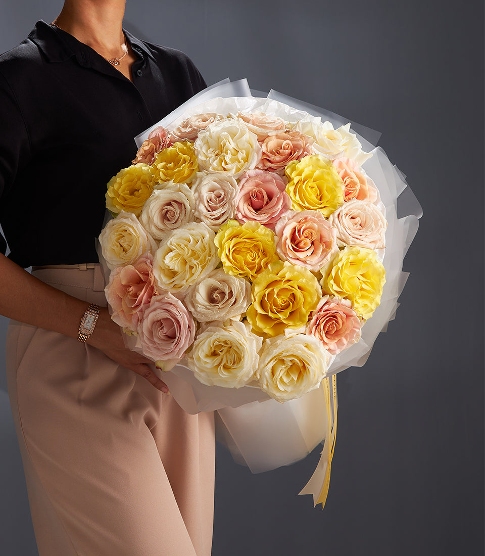 Large round bouquet of yellow, cream, and pink roses with water droplets, wrapped in white paper and yellow ribbon