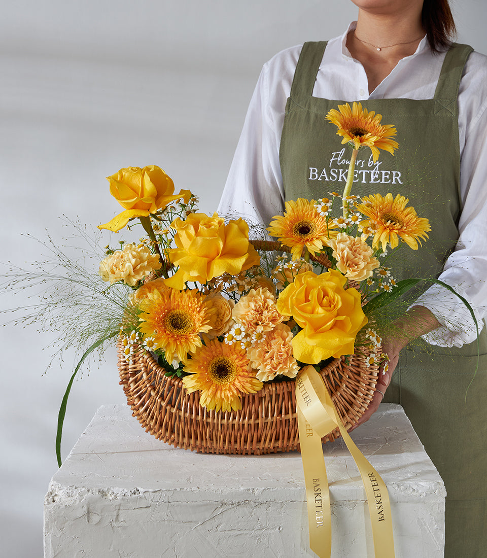 Florist holding golden yellow rose and gerbera daisy basket with orange carnations and ribbon, birthday gift arrangement