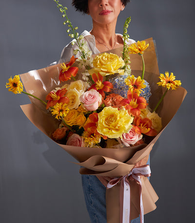 Vibrant bouquet of yellow roses, orange lilies, and pink roses with blue hydrangea wrapped in brown paper and ribbon