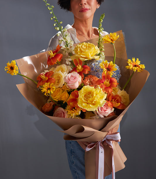 Vibrant bouquet of yellow roses, orange lilies, and pink roses with blue hydrangea wrapped in brown paper and ribbon