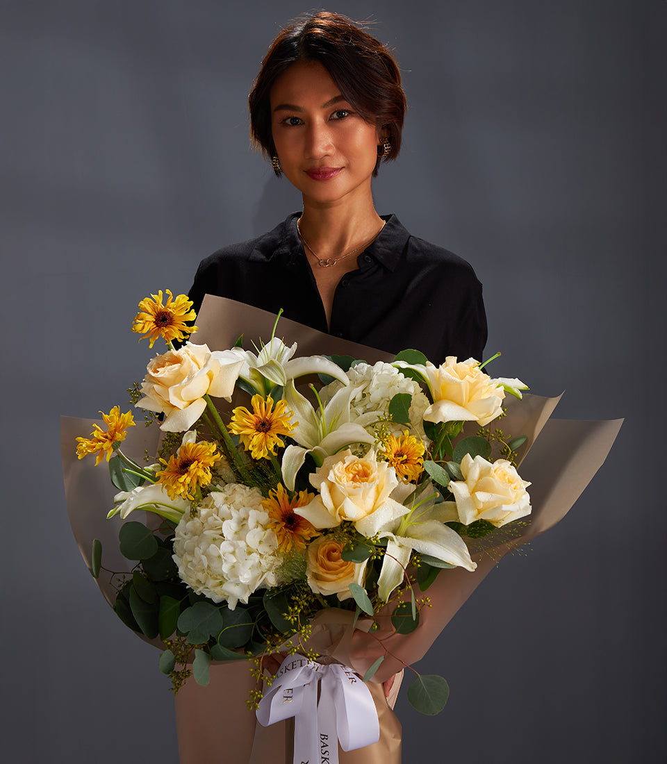 Woman holding large bouquet of yellow roses, white lilies, hydrangeas, and yellow-orange flowers with eucalyptus in light