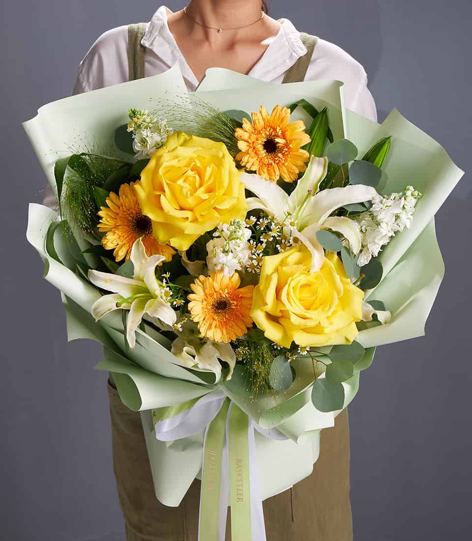 Vibrant yellow roses, white lilies, and orange gerberas in light green wrapping with eucalyptus foliage and white ribbon