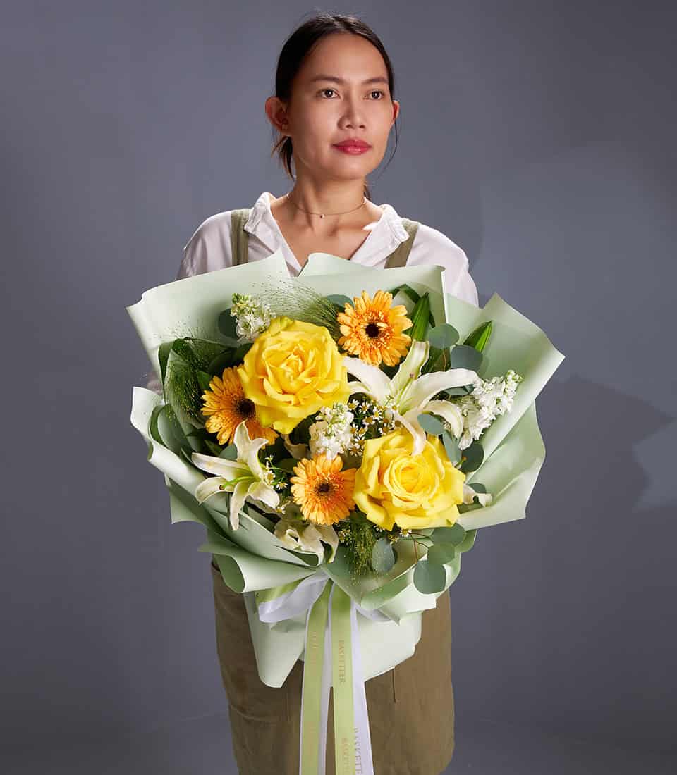 Vibrant yellow roses, white lilies, and orange gerbera bouquet with green foliage in light green wrap