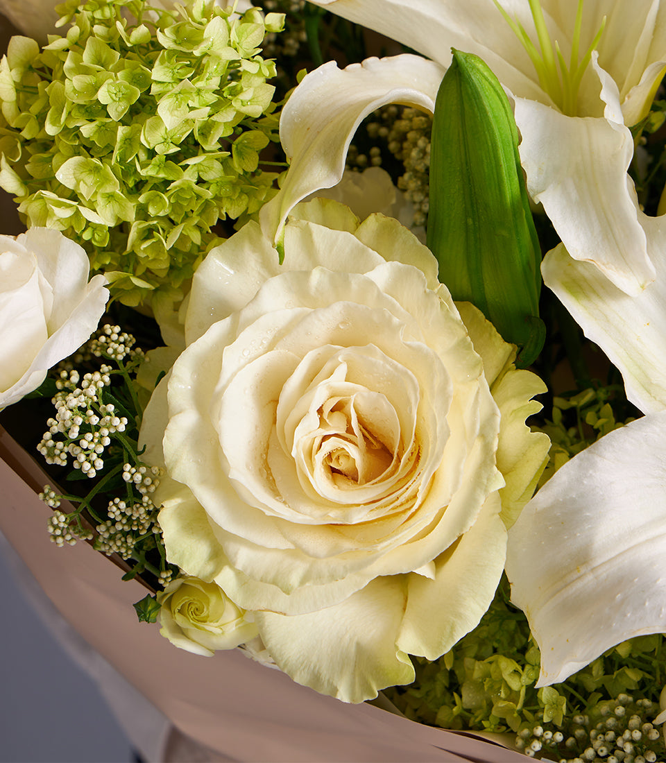 Creamy white rose and lily bouquet with fresh green foliage, wrapped in blush paper