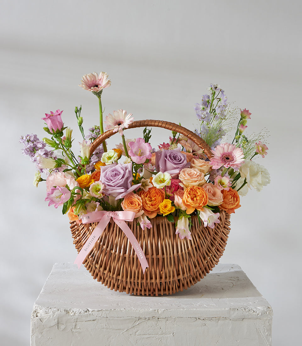 Lavender roses, peach spray roses, and pink daisy bouquet in wicker gift basket with pink ribbon