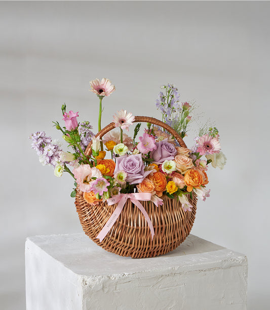 Lavender, peach spray roses and pink daisies in woven wicker basket with pink bow