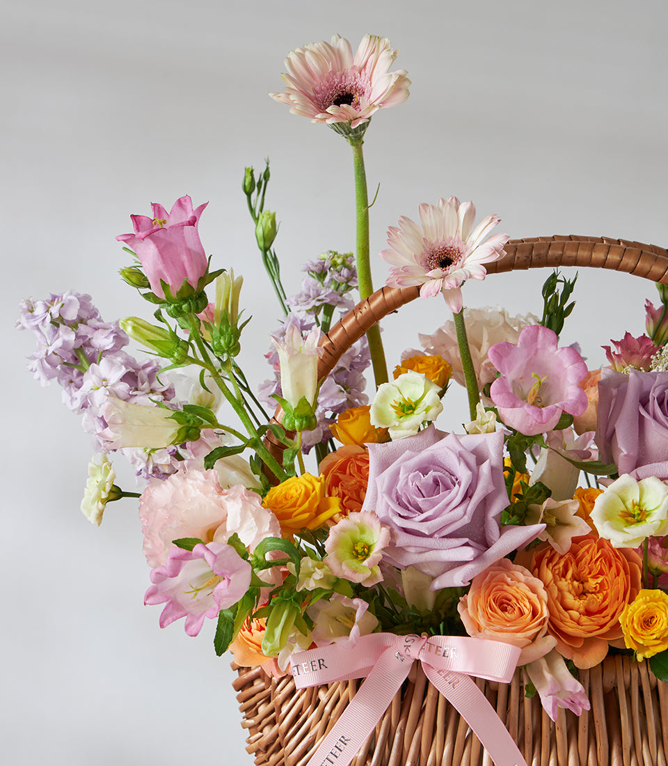 Lavender roses, peach spray roses, pink daisies in woven wicker basket with pink ribbon