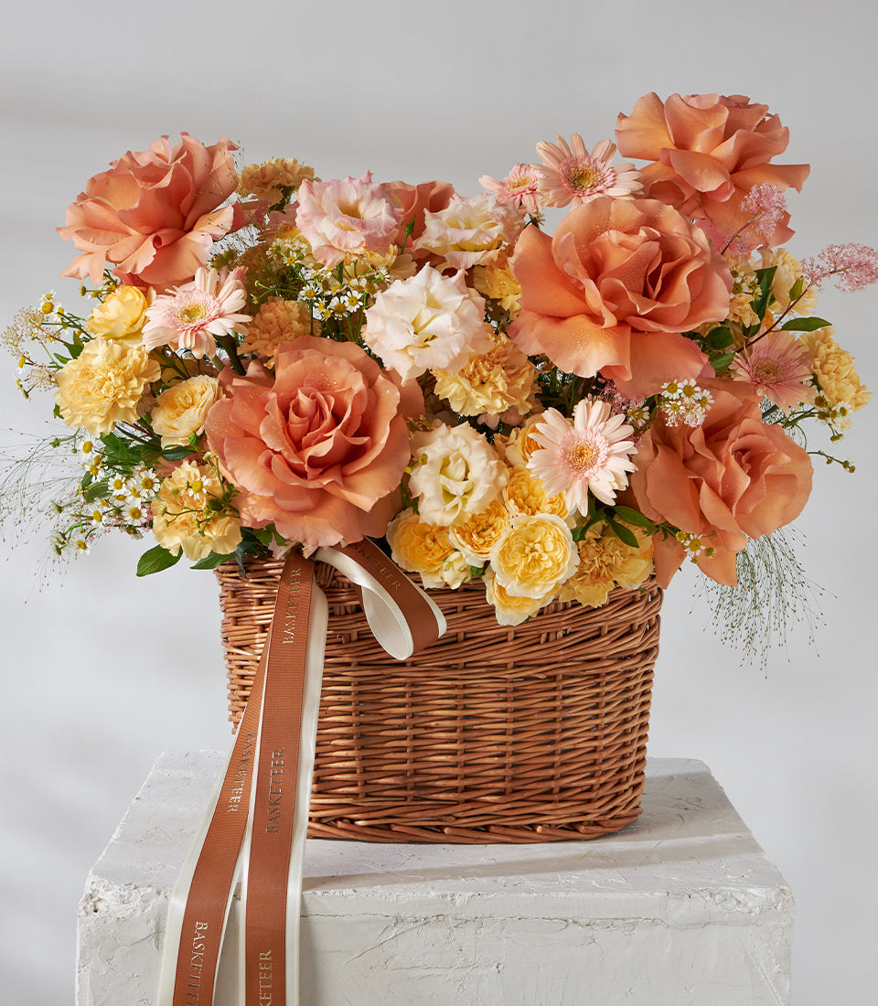 Vibrant orange and peach roses, carnations, and chrysanthemums in woven wicker basket with white ribbon
