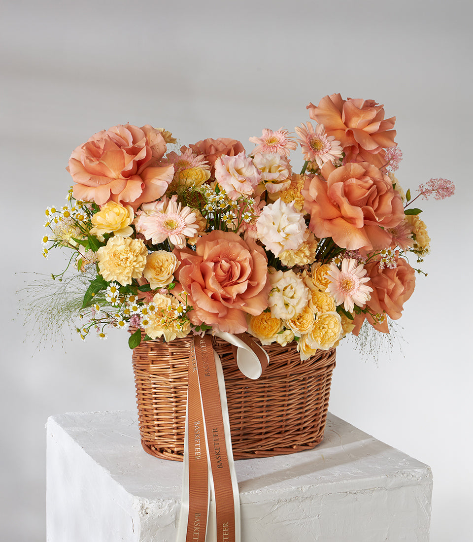 Vibrant orange and peach roses, carnations, and chrysanthemums in woven wicker basket with ribbon