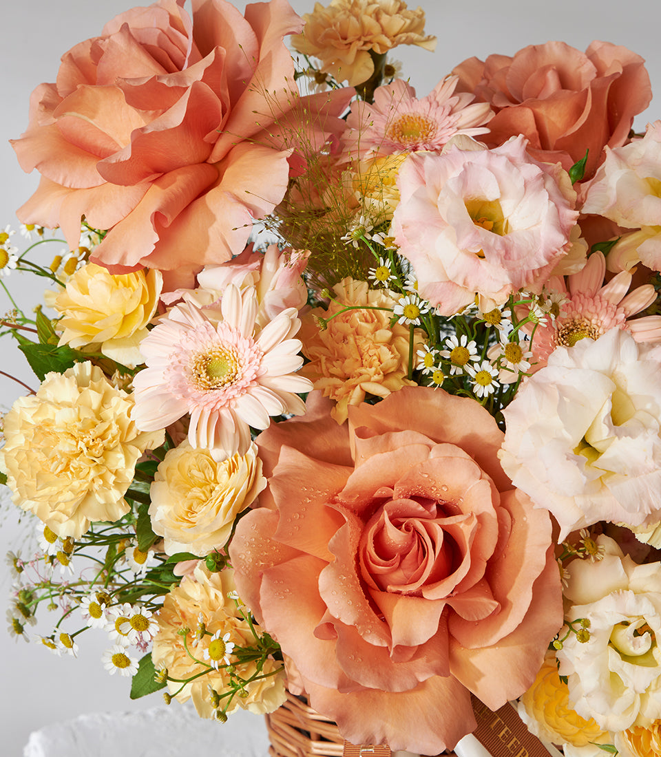 Peach roses, pink gerberas, and yellow carnations in woven wicker basket with green foliage